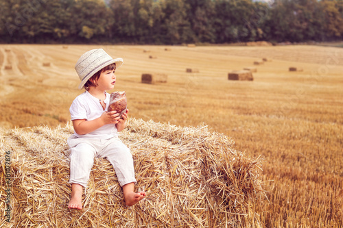 Cute little boy in straw hat eating bread sitting on hay stack in harvested yellow wheat field. Summer lifestyle. Child nutrition. Rural scene. Copy space © Tetiana Soares