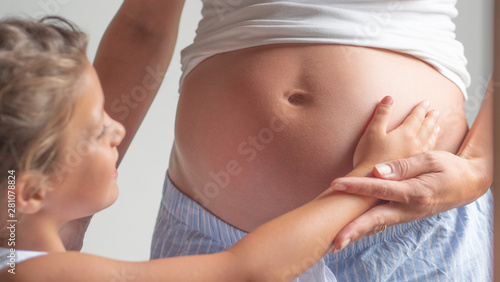 Authentic shot of a lovely little daughter is touching mother's pregnant belly, she is feeling baby at mother tummy and awaiting the birth of her little brother or sister.