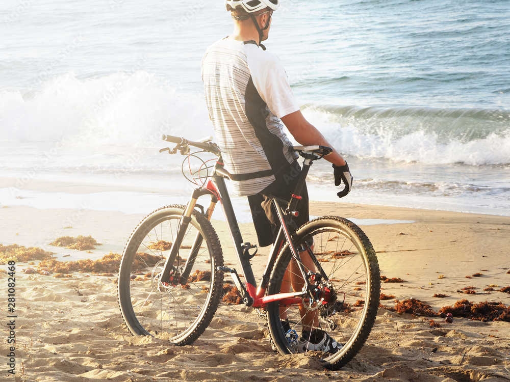 Fototapeta premium Cyclist resting on the beach of the Atlantic Ocean. Back view. Evening sunset. sun flare.