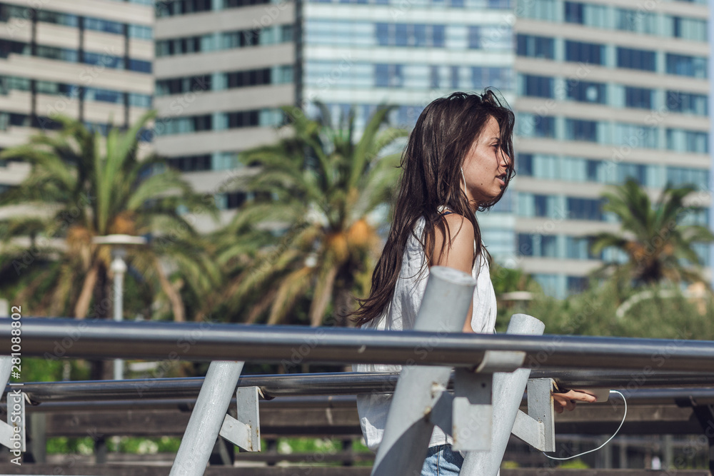 Beautiful young woman walking around the city and listening to the music on the phone. Portrait of gorgeous brunette girl enjoying the music over summer background with palm trees and buildings. Beautiful young woman walking around the city and listening to the music on the phone. Portrait of gorgeous brunette girl enjoying the music over summer background with palm trees and buildings.