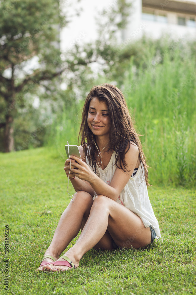 Portrait of cheerful brunette girl sitting on the garss in summer park. Close-up photo of young beautiful woman chatting and listening to music on her smartphone outdoors. Portrait of cheerful brunette girl sitting on the garss in summer park. Close-up photo of young beautiful woman chatting and listening to music on her smartphone outdoors.