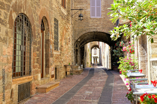 Fototapeta Naklejka Na Ścianę i Meble -  Beautiful arched street in the medieval old town of Assisi with flowers and restaurant tables, Italy