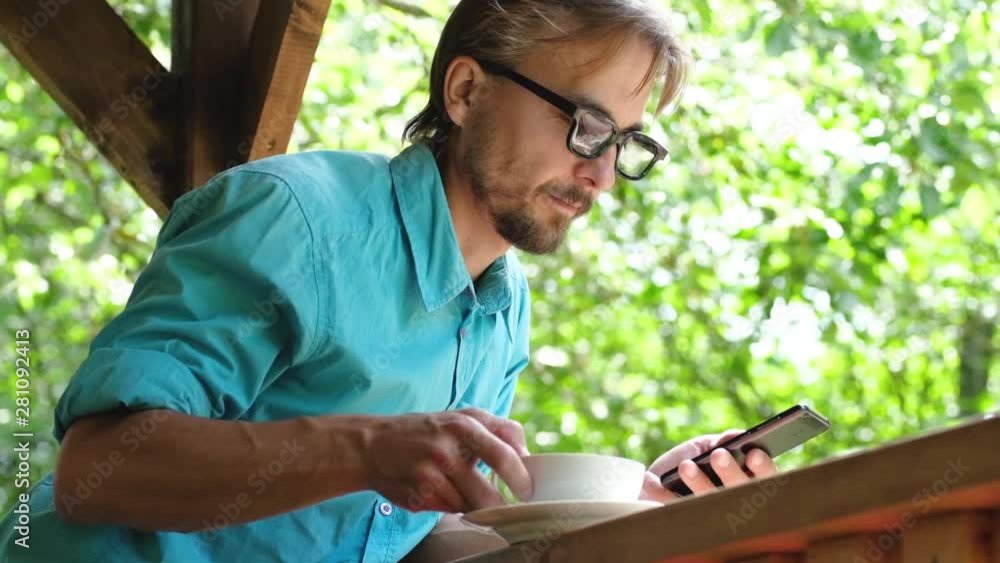 Handsome bearded man in casual wear with glasses drinking coffee and uses smartphone in summer open terrace cafe. Handsome bearded man in casual wear with glasses drinking coffee and uses smartphone in summer open terrace cafe.