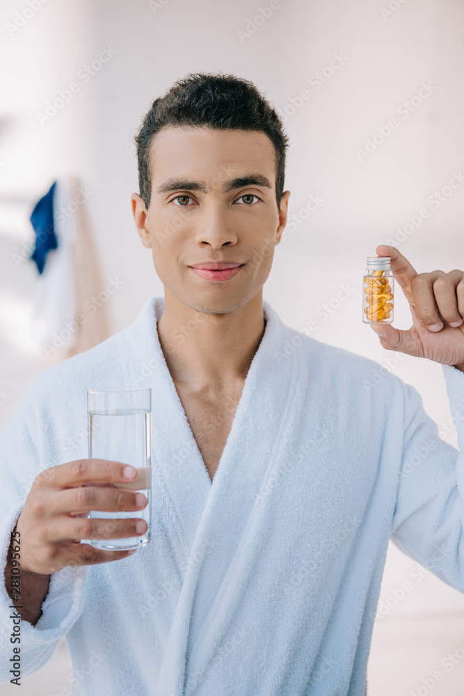 handsome man in bathrobe holding bottle with vitamins and glass with water and looking at camera handsome man in bathrobe holding bottle with vitamins and glass with water and looking at camera