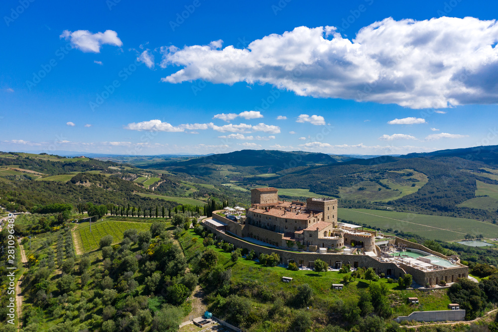 Fototapeta premium A castle stands on top of a Tuscan hill surrounded by cypress trees. Mountains shape the background landscape.