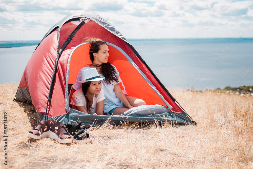 Two sisters are resting in a campsite in nature. Two young women in a tent on a background of a lake Two sisters are resting in a campsite in nature. Two young women in a tent on a background of a lake