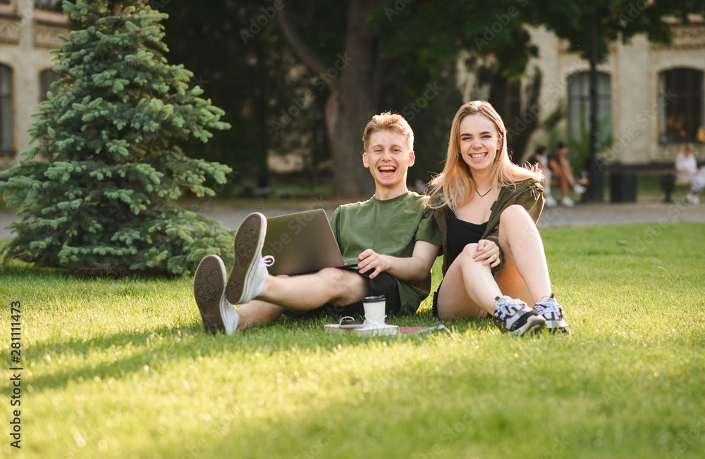 Portrait of two laughing attractive young students using laptop and sitting on grass in college park. Couple of cheerful students in university garden feeling careless. Friendship, education concept. Portrait of two laughing attractive young students using laptop and sitting on grass in college park. Couple of cheerful students in university garden feeling careless. Friendship, education concept.