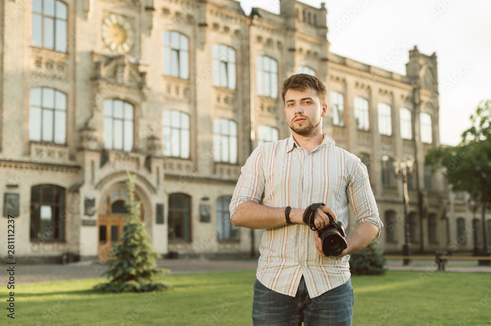 Professional male photographer with a beard holding camera standing on the architecture background in the park looking at camera. Handsome young man with camera is satisfied working as a photographer.