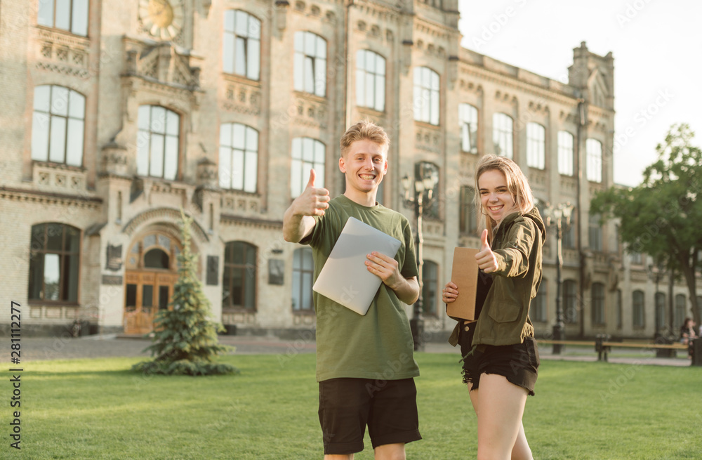 Two cheerful students with broad smiles raising thumbs up feeling successful. Exams are over, good grades, cheerful students near the university in the park with laptop and notebook. Like concept.