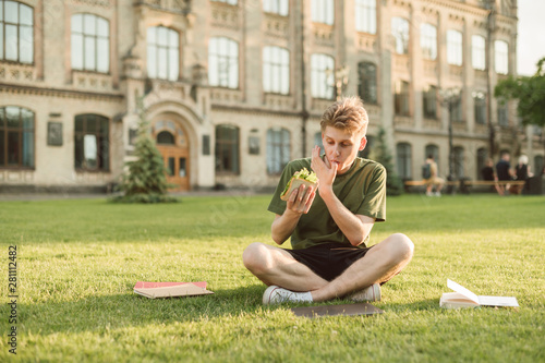 Attractive cute male student having lunch on the college lawn eating delicious sandwich licking his finger. Handsome university student eating appetizing sandwich having rest on a break near campus. © bodnarphoto