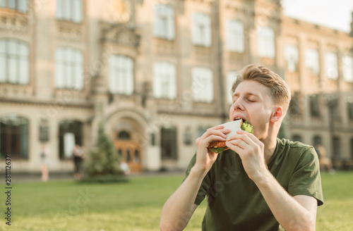 Close-up portrait of a young man sitting on the background of the building biting a sandwich with eyes closed.Hungry student eats an appetizing food of man on the background of a university building