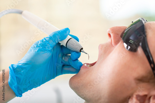 Young man visiting the dental office. Dentist hands in protective gloves using ultrasound to clean patient's teeth. Closeup of open mouth.