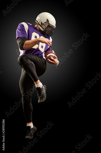 American football player holding a ball while jumping away from a strike. Black background, copy space. Violet with white sportswear. © Georgii