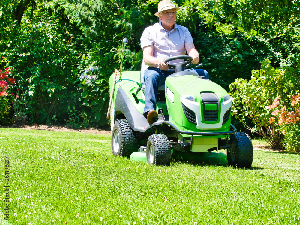 Fototapeta premium Senior man driving a tractor lawn mower in garden with flowers