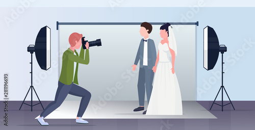 professional wedding photographer shooting on camera newly weds couple bride and groom embracing posing in modern photo studio interior full length flat horizontal