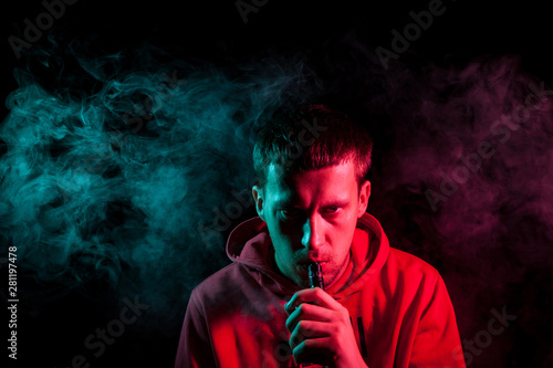 A young brunette man holds in his hand and drags on an electronic cigarette with a vape while smoking and releases smoke to the sides, his face is highlighted with red and green light.