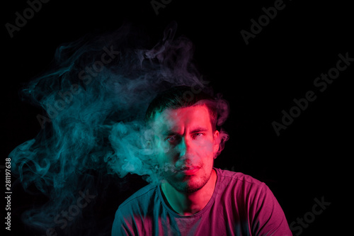 Close up portrait of the face of an adult serious man exhales green toxic smoke while smoking e-cigarette and vape illuminated with blue and red colored light on a black background. Harm to health.