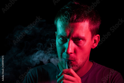 A young brunet male turned his head to the side, holds in his hand and inhales e-cigarette with a vape while smoking and releases smoke to the side, his face is highlighted with red and green light