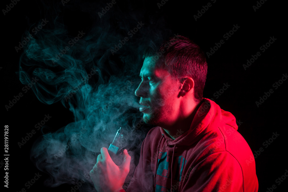 Close up portrait of the face of an adult serious man exhales green toxic smoke while smoking e-cigarette and vape illuminated with blue and red colored light on a black background. Harm to health. Close up portrait of the face of an adult serious man exhales green toxic smoke while smoking e-cigarette and vape illuminated with blue and red colored light on a black background. Harm to health.