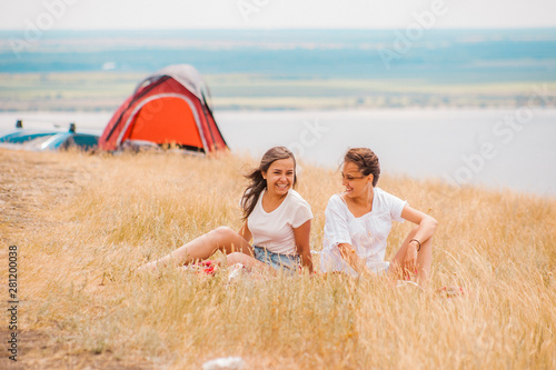 In the foreground, two friends are sitting on a hill by the river in the grass, talking on a summer vacation, in the background there is a tent for relaxing next to the car