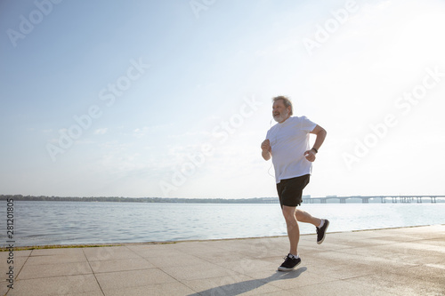 Senior man as runner with armband or fitness tracker at the riverside. Caucasian male model practicing jogging and cardio trainings in summer's morning. Healthy lifestyle, sport, activity concept.