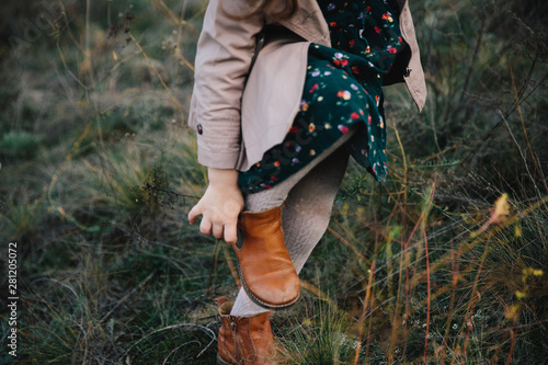 fair-haired girl in a beige coat shoes rubber boots on the dry grass and autumn trees background.