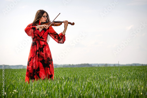 Young woman in red dress playing violin in green meadow - image