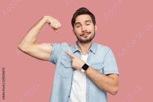 I am strong and I can do anything. Portrait of proud handsome bearded young man in blue casual shirt standing pointing and showing his strong bicep