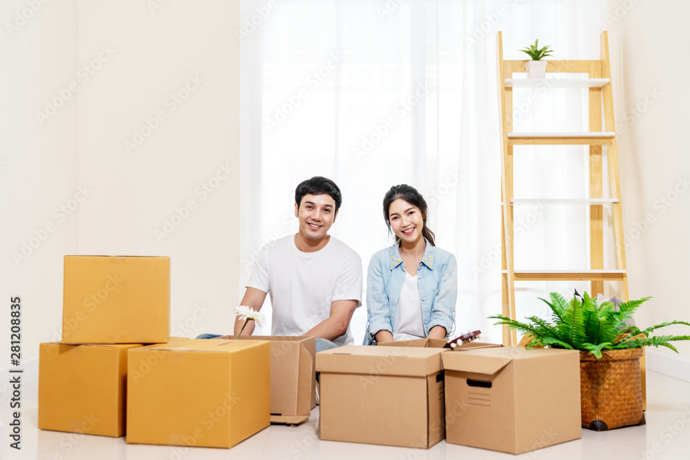 Portrait of young attractive happy asian couple man and woman sitting at new home smiling to camera with carton package box storage to move in empty house. Young married asian relocating home concept.