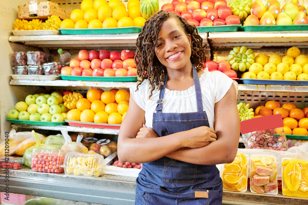Portrait of African young woman in jeans apron standing with arms crossed and smiling at camera with shelves of fruits and vegetables in the background