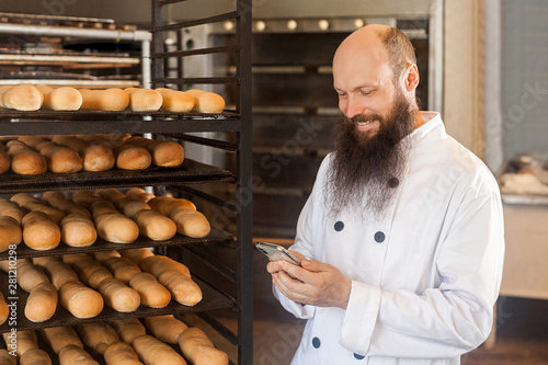 Portrait of satisfied young adult businessman baker with long beard in white uniform standing in bakery and have online order by phone, using phone with toothy smile Indoor, profession concept