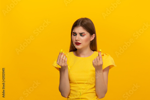 Young model in a yellow tee having problems listening to music with wired headphones standing over yellow background.