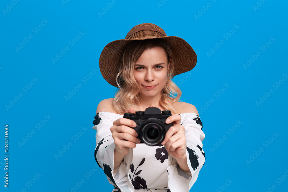 Portrait of a model with curly blond hair in a sundown hat and white dress takes photos with retro camera standing on a blue background. Portrait of a model with curly blond hair in a sundown hat and white dress takes photos with retro camera standing on a blue background.