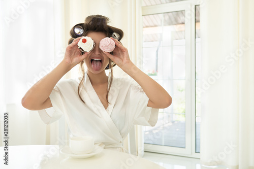 beautiful young brunette woman in hair curlers showing tongue holding two cupcakes in both hands near her eyes sitting with cup of coffee in bright kitchen