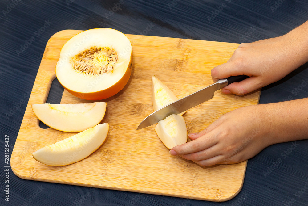 Half a yellow melon and slices. Women's hands cut melon on a cutting Board, on a black wooden background.