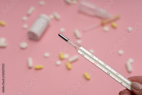 Close-up. Thermometer in female hand on the background of tablets, pills and other medicine scattered on a pink background. Treatment of illness, malaise.