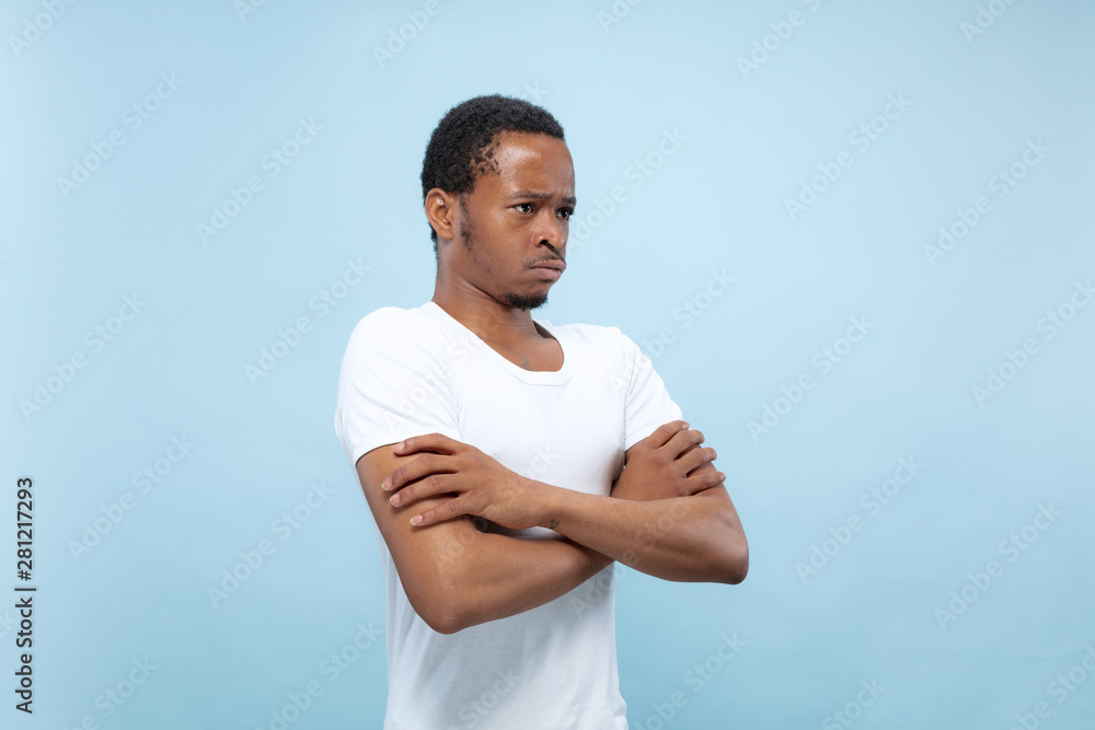 Half-length close up portrait of young african-american man in white shirt on blue background. Human emotions, facial expression, ad, sales, concept. Standing hands crossed and sad, upset or calm. Half-length close up portrait of young african-american man in white shirt on blue background. Human emotions, facial expression, ad, sales, concept. Standing hands crossed and sad, upset or calm.