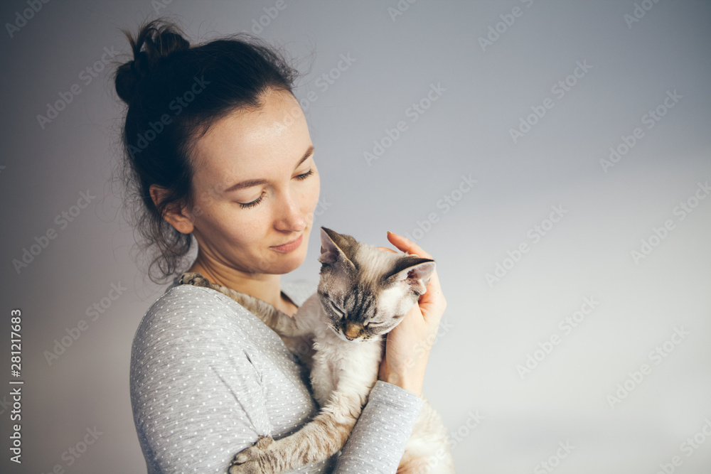 Woman is holding and cuddling a little point color Devon Rex cat. Kitten is felling relaxed and content and purrs. Selective focus