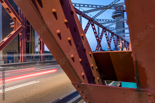 The red Spijkenisser Bridge at dusk seen through a steel pillar with trail lights from fast moving traffic and the bridge control tower at the background photo