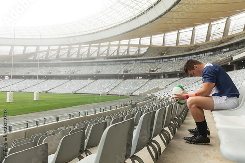 Upset Caucasian male rugby player sitting with rugby ball in stadium