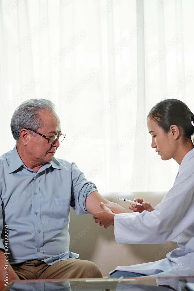 Young nurse in white coat sitting on sofa and make an injection in hand to her senior patient at home Young nurse in white coat sitting on sofa and make an injection in hand to her senior patient at home