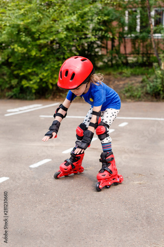 A small child in bright clothes, in a red helmet and protection goes on the road on rollers. Children's sport, active leisure.