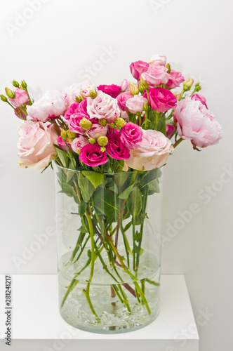 Bouquet of pink roses  peony flowers in glass vase on a shelf against white wall background.