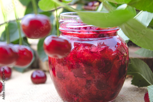 Cherry jam in a glass jar on a background of green leaves on a table of wooden boards.