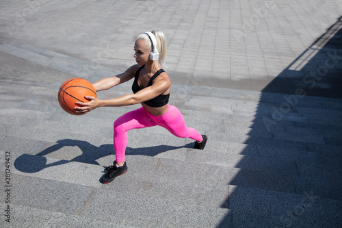 A young athletic woman in shirt and white headphones working out listening to the music at the street outdoors. Doing lunges with the ball. Concept of healthy lifestyle, sport, activity, weight loss.