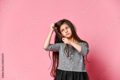 little brunette girl with long hair holds a strand of her hair and looks at it. Hair care and haircut.