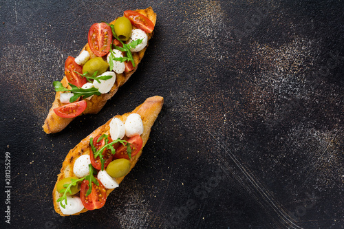 Caprese bruschetta toasts with cherry tomatoes, mozzarella, olives and basil on old dark background. Top view.