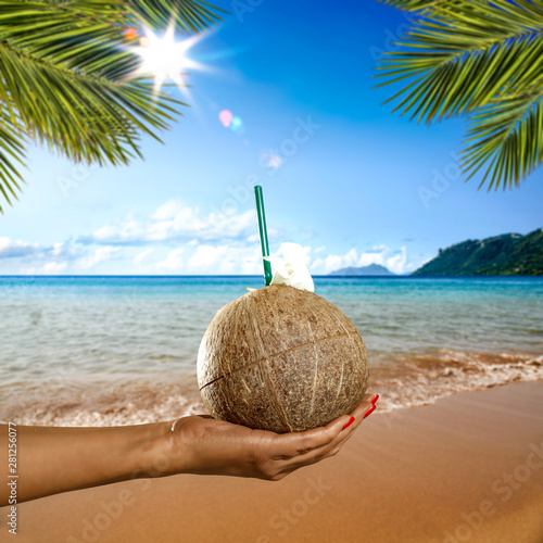 A coconut with a straw holding in a hand on a sunny beach view.