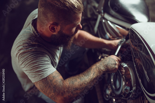 Handsome bearded man in leather jacket and sun glasses is sitting on the motorcycle in the repair shop
