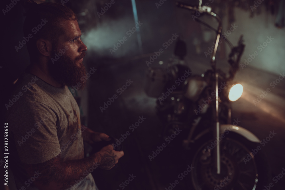 Handsome brutal man with a beard is standing in his garage against the background of a motorcycle and looking away